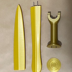 May include: A gold-colored plastic sword model kit with four pieces: a blade, a hilt, a stand, and a base. The pieces are arranged on a brown surface.