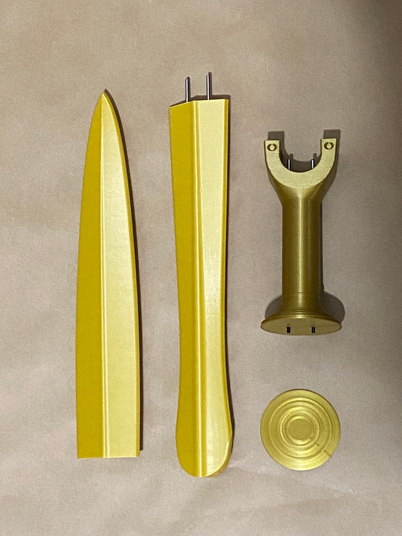 May include: A gold-colored plastic sword model kit with four pieces: a blade, a hilt, a stand, and a base. The pieces are arranged on a brown surface.