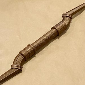 May include: A brown, elongated, curved object with a central cylindrical grip featuring a geometric, repeating pattern. The object has raised, decorative bands at both ends of the grip, with additional intricate, embossed floral-like detailing.