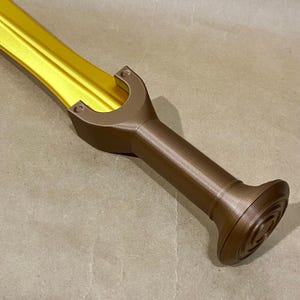 May include: A toy sword featuring a bright metallic gold blade and a dark brown handle. The hilt has a U-shaped guard that connects to a cylindrical grip, ending in a circular pommel with concentric ring details visible on the surface.