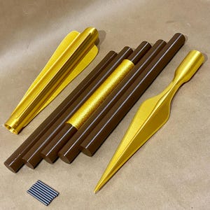 May include: A set of gold-colored spear parts, including a spearhead, shaft sections, and a tip. The shaft sections are brown and have a textured surface. There are also small metal pins included.