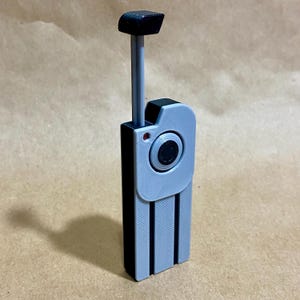 May include: A 3D-printed device with a rectangular black base and a light gray top section. It features a circular lens and a small red dot. A black rectangular handle extends upwards from the top.