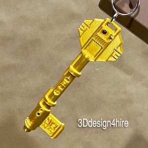 May include: A gold-colored key with a detailed design. The key has a large, ornate head with a red dot in the center. The key is attached to a silver keyring.