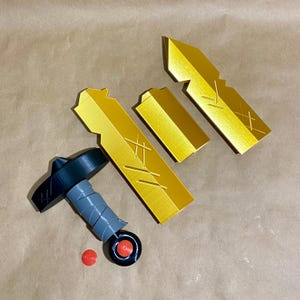 May include: A set of five gold-colored plastic pieces that can be assembled to create a sword. The pieces include a handle, a blade, and three smaller pieces. The handle is black and gray and has a small red button on the side.