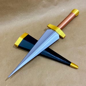 May include: A decorative dagger with a silver blade, a copper-colored handle, and gold-colored accents. The dagger is resting on top of a black sheath with gold trim. The overall design is symmetrical and detailed.