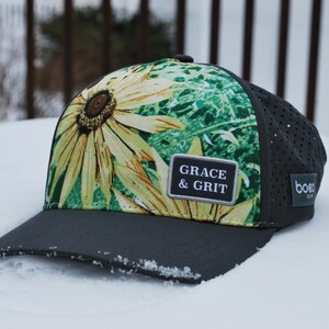 Peut inclure: Une casquette de baseball noire et verte avec un imprimé floral et un patch qui dit "GRACE & GRIT". La casquette est posée sur une surface blanche.