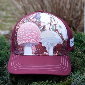 May include: A maroon trucker hat with a white mesh back. The front of the hat features a colorful print of two mushrooms in a forest setting.