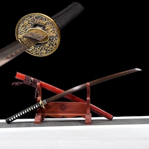 May include: A traditional Japanese katana sword with a black scabbard and a red and black handle. The sword is displayed on a wooden stand with a red and gold design.