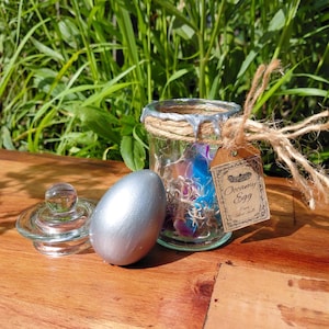 May include: A silver-painted egg sits beside a small glass jar with a lid. The jar is wrapped in twine and contains colourful feathers. A brown paper tag attached to the twine reads Occamy Egg Pure Silver Shells in black decorative text.