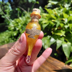 Könnte beinhalten: Ein kleines, tropfenförmiges Glasfläschchen gefüllt mit einer schimmernden goldenen Flüssigkeit. Die Flasche ist mit einem Korken verschlossen und trägt ein beigefarbenes Papieretikett mit dem in schwarzer Schrift gedruckten Text Felix Felicis.