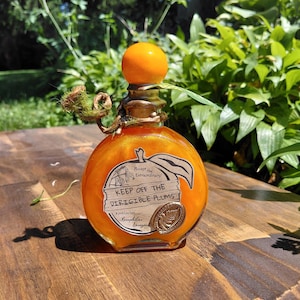 May include: A round glass bottle filled with shimmering orange liquid, topped with a bright orange spherical stopper. A label shaped like a plum features the text "KEEP OFF THE DIRIGIBLE PLUMS" and "Accept the Extraordinary," with a small metallic fern seal.