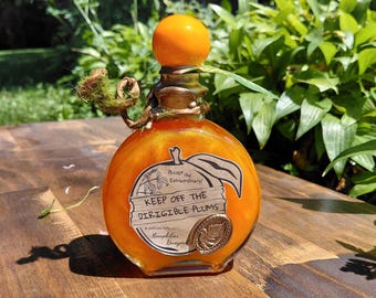 Könnte beinhalten: Eine runde Glasflasche gefüllt mit schimmernder orangefarbener Flüssigkeit, gekrönt von einem leuchtend orangefarbenen kugelförmigen Verschluss. Ein pflaumenförmiges Etikett trägt den Text "KEEP OFF THE DIRIGIBLE PLUMS" und "Accept the Extraordinary".