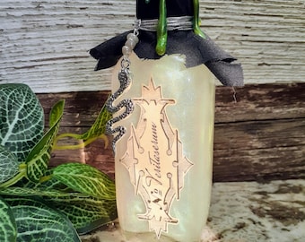 Könnte beinhalten: Eine dekorative Glasflasche mit schimmerndem Finish, mit einem schwarzen Deckel mit grünen Tropfen. Ein silberner Schlangenanhänger hängt an der Flasche. Auf dem Etikett steht "Veritaserum".