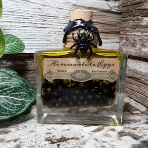 Puede incluir: Una botella cuadrada de vidrio llena de líquido amarillo y esferas negras, etiquetada como "Acromantula Eggs". La botella está sellada con un corcho y cera negra, adornada con una araña negra. La etiqueta dice "Glass A" y "Non-Tradeable".