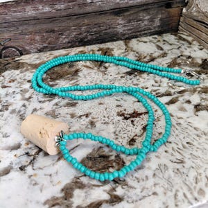 May include: A turquoise beaded necklace with a natural cork pendant. The necklace features small, round beads and a silver-colored clasp. The cork pendant is cylindrical and has a silver-colored floral accent.