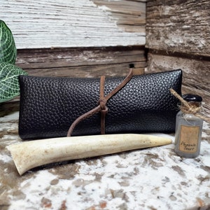 Puede incluir: Una bolsa de cuero negro con un lazo de cuero marrón, una cuerna de ciervo blanca y una pequeña botella de vidrio con una etiqueta que dice "Phoenix Tears".