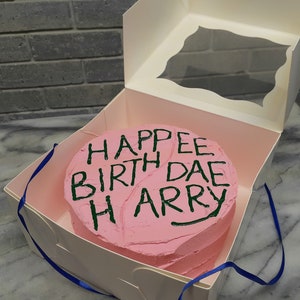 Peut inclure: Un gâteau rond au glaçage rose dans une boîte en carton blanc avec une fenêtre transparente. Le dessus du gâteau présente une inscription en glaçage vert foncé HAPPEE BIRTH DAE HARRY, écrite dans un style manuscrit, avec un ruban bleu visible.