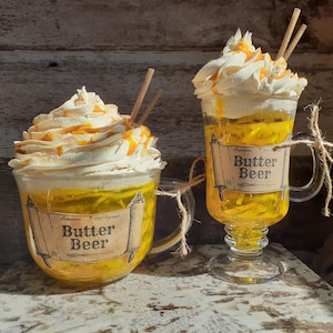 May include: Two clear glass mugs of Butter Beer. The mugs are filled with a yellow liquid and topped with whipped cream and caramel drizzle. Each mug has a label that reads "Butter Beer". The mugs are tied with twine.