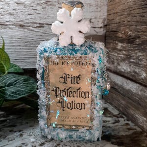 May include: A decorative potion bottle with a snowflake stopper. The label reads "Fire Protection Potion" and "Protects Against Magical Fires." The bottle is embellished with blue and white accents, and a small chain with blue beads.