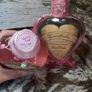 May include: Two heart-shaped glass bottles filled with pink liquid. One bottle has a pink wax seal with a rose design. The other bottle has a label that reads "Amortentia Love Potion". Both bottles are decorated with pink accents.