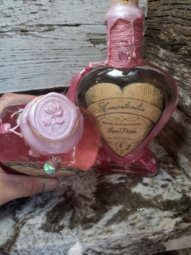 May include: Two heart-shaped glass bottles filled with pink liquid. One bottle has a pink wax seal with a rose design. The other bottle has a label that reads "Amortentia Love Potion". Both bottles are decorated with pink accents.