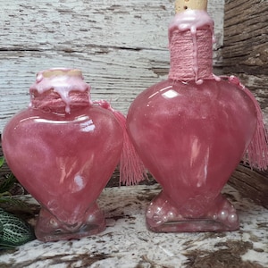 May include: Two heart-shaped glass bottles filled with a shimmering pink liquid. The bottles have a cork stopper, wrapped in pink twine, and adorned with pink tassels. The bottles are on a wooden background.