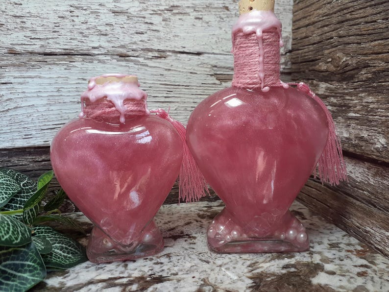 May include: Two heart-shaped glass bottles filled with a shimmering pink liquid. The bottles have a cork stopper, wrapped in pink twine, and adorned with pink tassels. The bottles are on a wooden background.