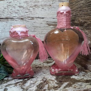 May include: Two heart-shaped glass bottles with pink accents. The bottles have a pink wax seal around the neck and a pink tassel. The bottles are on a marble surface with a wooden background.