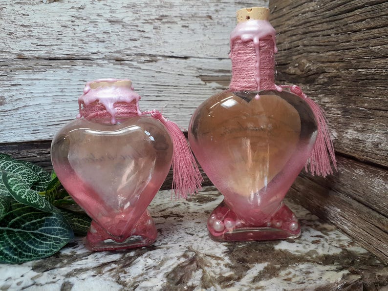 May include: Two heart-shaped glass bottles with pink accents. The bottles have a pink wax seal around the neck and a pink tassel. The bottles are on a marble surface with a wooden background.