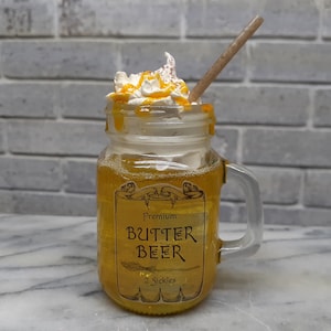 May include: A mason jar filled with a golden-colored beverage, topped with whipped cream and a drizzle of caramel. A brown straw is inserted into the drink. The jar has a handle and a label that reads "Premium Butter Beer".