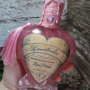May include: A heart-shaped pink glass bottle labeled "Amortentia Love Potion." The bottle is decorated with pink twine, a pink tassel, and a heart-shaped charm. The label includes the text "Caution: World's Most Powerful" and "Causes intense Infatuation & obsession."