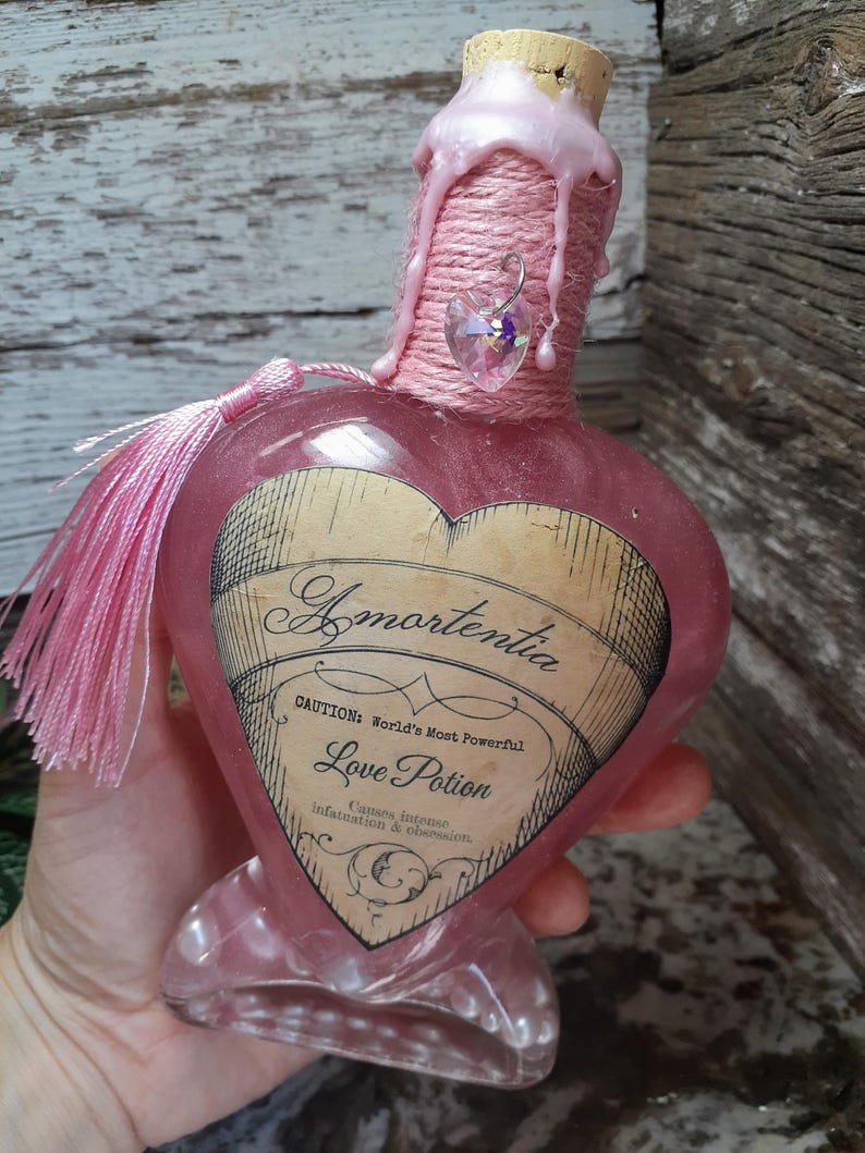 May include: A heart-shaped pink glass bottle labeled "Amortentia Love Potion." The bottle is decorated with pink twine, a pink tassel, and a heart-shaped charm. The label includes the text "Caution: World's Most Powerful" and "Causes intense Infatuation & obsession."