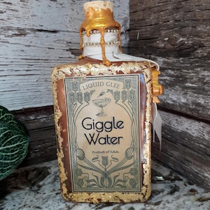 以下が含まれることがあります： 「Liquid Glee Giggle Water, Product of U.S.A.」とラベルされた装飾ボトル。 ボトルは金色のトップとアクセントがあり、白いリボンとラインストーンのトリムが付いています。 ラベルはアールヌーボーのデザインです。