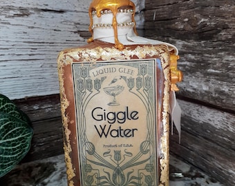 Könnte beinhalten: Eine dekorative Flasche mit der Aufschrift "Liquid Glee Giggle Water, Product of U.S.A.". Die Flasche hat einen goldfarbenen Deckel und Akzente, mit einem weißen Band und Strassbesatz. Das Etikett hat ein Jugendstil-Design.
