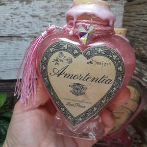 May include: A heart-shaped pink glass bottle labeled "Amortentia" with a decorative label. The bottle has a pink wax seal, a crystal charm, and a pink tassel. The text "Caution: World's Most Powerful Love Potion" is also visible.