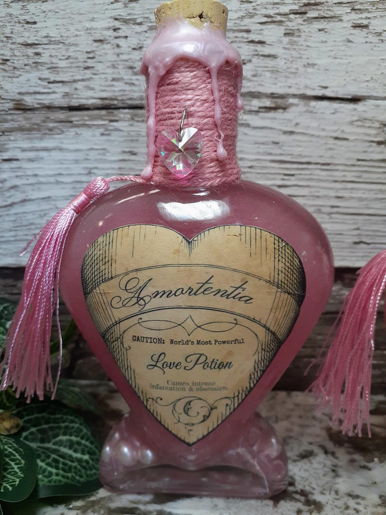 May include: A pink, heart-shaped glass bottle labeled "Amortentia Love Potion." The bottle has a cork stopper, pink wax, and a pink tassel. A heart-shaped crystal hangs from the neck. The label reads "Causes intense infatuation & obsession."