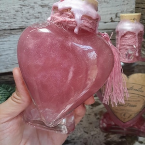 May include: A heart-shaped glass bottle filled with pink liquid, adorned with a pink rope and tassel. The bottle's top is sealed with a cork and pink wax. A smaller bottle with a similar design is in the background.