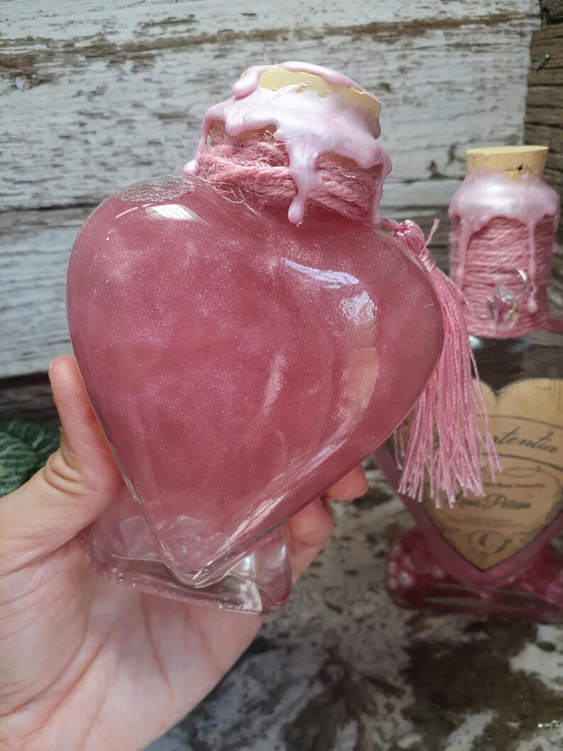 May include: A heart-shaped glass bottle filled with pink liquid, adorned with a pink rope and tassel. The bottle's top is sealed with a cork and pink wax. A smaller bottle with a similar design is in the background.