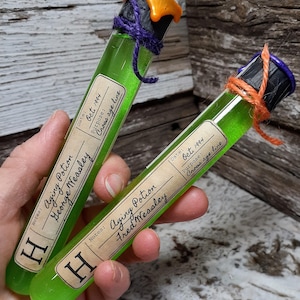 Puede incluir: Dos tubos de ensayo de vidrio llenos de líquido verde y etiquetados "Poción de envejecimiento" con los nombres "George Weasley" y "Fred Weasley". Las etiquetas también incluyen la fecha "Oct. 1994" y el propósito "Cruzar la línea de edad".