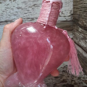 May include: A heart-shaped, pink glass bottle filled with a shimmering liquid. The bottle has a clear base and is topped with a cork and pink wax. The neck is wrapped with pink twine and a pink tassel hangs from the side.
