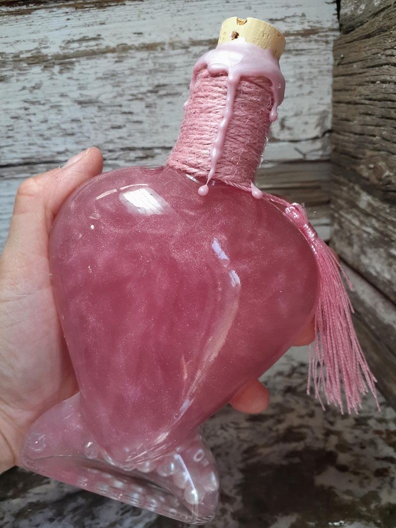 May include: A heart-shaped, pink glass bottle filled with a shimmering liquid. The bottle has a clear base and is topped with a cork and pink wax. The neck is wrapped with pink twine and a pink tassel hangs from the side.