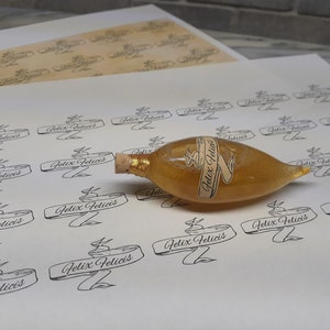May include: A sheet of paper with multiple printed labels that say "Felix Felicis" in a cursive font. A small, clear glass bottle with a gold liquid and a cork stopper is sitting on the paper. The bottle has a label that says "Felix Felicis" in a cursive font.