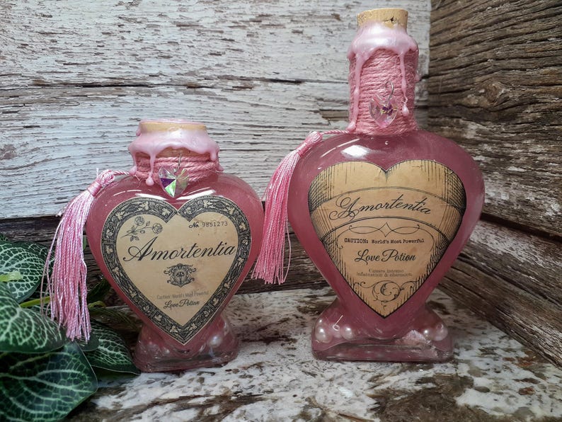 May include: Two heart-shaped, pink glass bottles labeled "Amortentia Love Potion." Each bottle has a cork stopper, pink string, and a tassel. The labels are vintage-style with decorative borders and text.