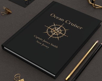 May include: A black journal with gold lettering that reads "Ocean Cruiser" and "Captain James Smith, New Jersey." A compass rose design is centered below the title. Gold pencils and binder clips are also visible.
