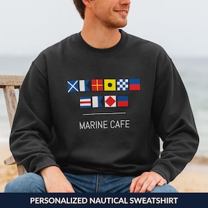 Op de afbeelding: Zwart sweatshirt met nautische vlagontwerpen en de tekst "MARINE CAFE". De sweatshirt heeft een ronde hals en lange mouwen. De onderkant van de afbeelding luidt "PERSONALIZED NAUTICAL SWEATSHIRT".