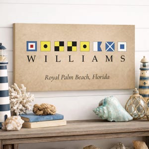 Op de afbeelding: Een rechthoekige canvas print met nautische vlaggen die "WILLIAMS" spellen boven "Royal Palm Beach, Florida". De print heeft een bruine achtergrond en wordt weergegeven op een houten plank met nautische decoratie.