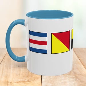 Peut inclure: Mug en céramique blanche avec une anse et un intérieur bleus. Le mug présente trois drapeaux de signalisation nautique bleus, rouges et jaunes. Le mug est posé sur une surface en bois.