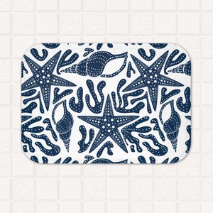 May include: A white bath mat with a navy blue pattern of starfish, seashells, and seaweed. The pattern is repeated throughout the mat.