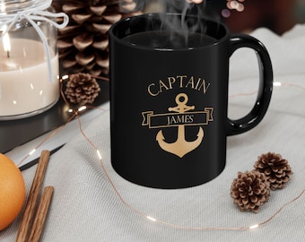 May include: A black ceramic mug with the words "CAPTAIN JAMES" and a gold anchor design. The mug is filled with a dark liquid, and steam rises from the top. The mug is on a light-colored surface with decorative elements.
