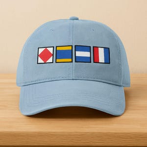 May include: A light blue baseball cap featuring embroidered nautical signal flags in red, yellow, blue, and white. The cap is made of a soft fabric and has a curved brim. The design is centered on the front of the cap.